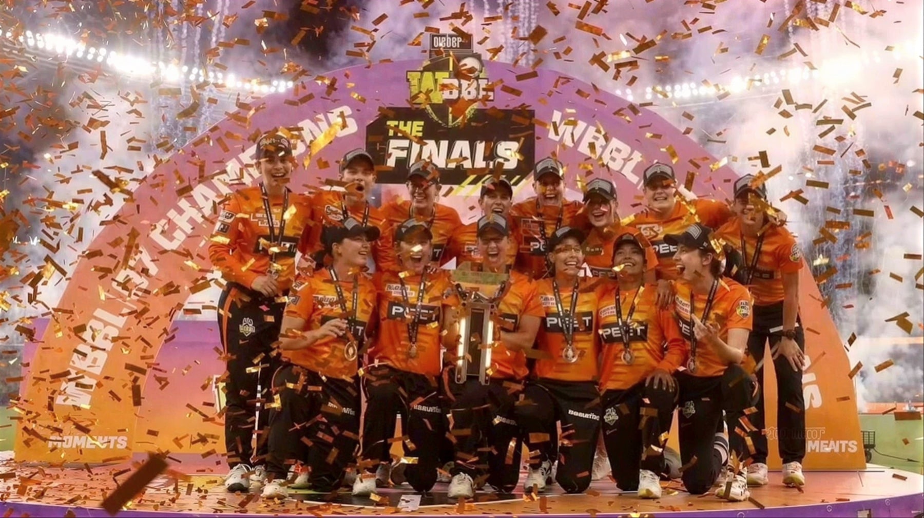 Perth Scorchers players celebrating together on stage with the WBBL trophy amid orange confetti and pyrotechnics
